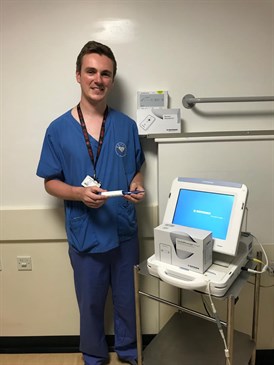 Adam Marzetti with the device. He is pictured wearing scrubs, holding the device and standing next to a computer-like machine