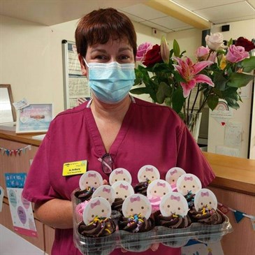 Jo Astbury with cakes celebrating vCreate's birthday. She is pictured wearing purple scrubs and holding a tray of chocolate cupcakes with icons of baby's faces on them. Behind her is a reception desk with flowers on.