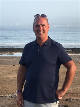 Paul Lloyd, spinal cord stimulator patient. He is pictured at the coast, with the sea behind him, wearing a navy polo shirt and light trousers. He has sunglasses on his head.