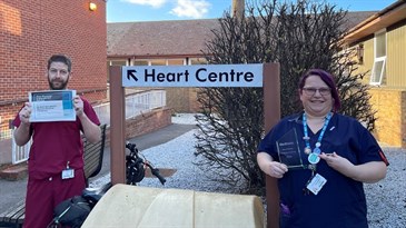 Niky Bradford and Craig Huckstepp with their awards for passing the training. They are pictured outside by a sign saying heart centre. Craig is on the left in maroon scrubs holding a certificate, Niky on the right in a blue uniform holding 