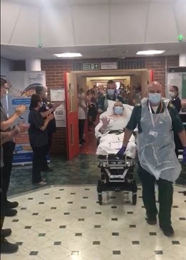 Alan Finch is wheeled out of QEQM. The picture shows him on a trolley being wheeled through the main reception area of the hospital, watched by staff and visitors who are clapping as he goes past.