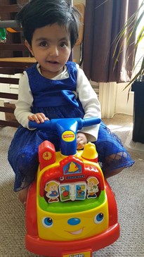 Amala now. She is pictured sitting on a ride-along toy wearing a blue dress.