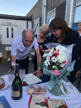 Anna and Alan Priestley sign the register after their wedding ceremony. Anna is wearing a blue trouser suit, Alan is in a white t-shirt and dark trousers. Alan is bent over signing a piece of paper, Anna is watching him as is the registrar. On the ta
