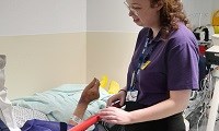 Avatar of Hannah Smith providing company and comfort to patients. Image shows her talking to a patient in a bed in ED. You can see the patient's hand and their body under a blanket with yellow falls socks on their feet. Hannah is wearing a volunteer'
