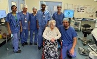 Avatar of Mary Teal with the surgical team who performed her cataract surgery. Image shows Mary in a wheelchair with a patch over one eye. The surgeon is kneeling next to her and the rest of the team are standing behind her. They are in an operating 