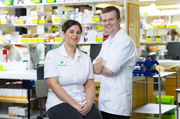 Ben Smith and Michelle Brown, pharmacy technicias who work in ED at the QEQM. They are pictured in a hospital pharmacy wearing white tunics. Michelle is sitting on a stool and Ben is standing next to her.