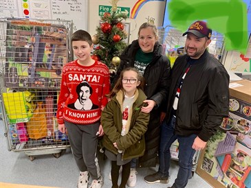 Betsy Boardman and her family with the presents she donated. Image shows Betsy and her older brother and her parents in front of a Christmas tree and a large container of toys. Everyone is wearing Christmas jumpers
