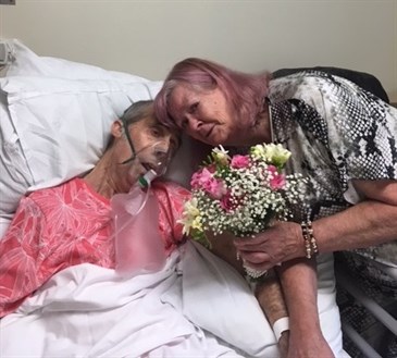 Bob and Anne Tomkins. Bob is lying in a hospital bed with an oxygen mask on. Anne is learning in to give him a cuddle, holding a bouquet. They married on the ward hours before he died.