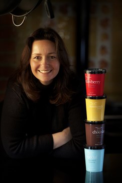 Chef Sally Newall, who was part of the Ashford Vineyard meals project. She is wearing a black top and pictured against a black background. Tubs of ice cream are stacked on top of each other next to her.