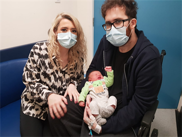 Cheryl Waters and Olly Toole with baby Lily. Olly is in a wheelchair, Cheryl is sitting beside him. He is holding Lily in his lap. Both the adults are wearing face masks.