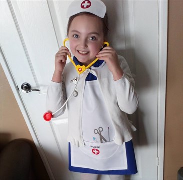 Reegan Price, who dressed as a nurse for superheroes day at school after NHS staff saved her dad's life. She is pictured wearing a nurse's uniform including hat, and holding a stethoscope, in front of a white door in her living room