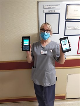 Debbie Delo with the audiobooks. She is pictured in a ward corridor holding an audio book device in each hand, wearing uniform and a face mask.
