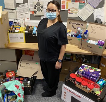 Elmas Deniz with some of the items that have been donated for the foodbank. She is in an office with boxes on the desk and floor full of donated items.