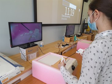 Fern Arnold tries some laporascopic surgery. Image shows a female student operaitng hand controls of instruments to reposition items in a box. We can see what is in the box on a screen in front of her.