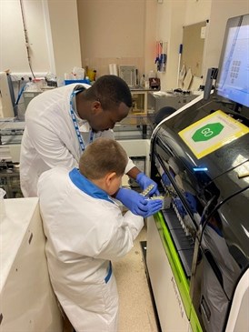 Finley Puddefoot loading blood samples. Image shows a young boy in a lab coat with a man in a lab coat by a machine in a hospital laboratory. They are loading samples into the machine.