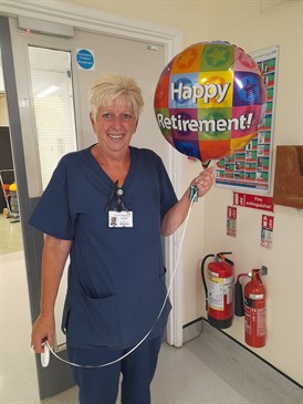 Gill Miller on her last day. Image shows her holding a balloon that says Happy Retirement. 