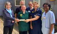 Avatar of Gill Young presenting the cheque to Leigh Carvill and the team. Image shows them standing in the garden area of the Cathedral Day Unit; Gill is on the left with five members of unit staff