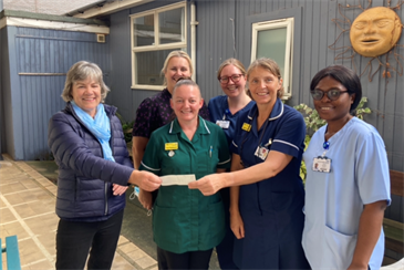 Avatar of Gill Young presenting the cheque to Leigh Carvill and the team. Image shows them standing in the garden area of the Cathedral Day Unit; Gill is on the left with five members of unit staff