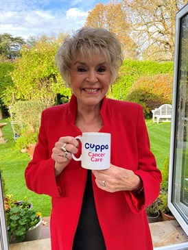 Gloria Hunniford is heading up the Cuppa for Cancer Care campaign. Image shows her holding a branded mug, with Cuppa for Cancer Care on, with a garden behind her