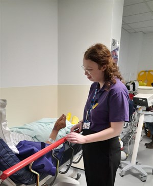 Hannah Smith providing company and comfort to patients. Image shows her talking to a patient in a bed in ED. You can see the patient's hand and their body under a blanket with yellow falls socks on their feet. Hannah is wearing a volunteer's t-shirt 