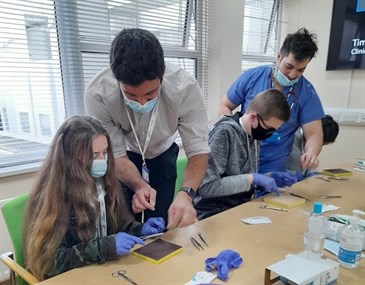Avatar of instruction in suturing techniques. Image shows two young people sitting at a table, with two older students watching on. The young people are attempting suturing stitches and are wearing blue surgical gloves. Everyone is wearing a face mas