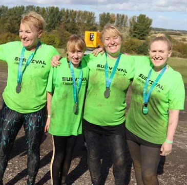 Jessie, second from left, with her family after the challenge. Image shows four women wearing green t-shirts. They are muddy and smiling at the camera with their arms around each other