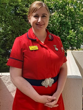 Katie Milner. Image shows Katie wearing a red nurses' uniform. She is pictured outside with trees behind her.