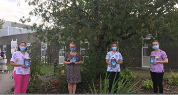 Author Loris Owen with staff from Padua Ward. They are outside the hospital with a tree behind them. The three staff members are wearing scrubs. All are holding a copy of the book and wearing a mask