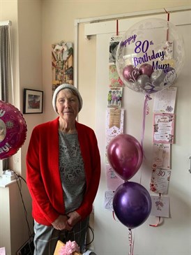 Margaret Smith on her birthday. She is pictured in her house, surrounded by balloons. She is wearing a red jacket and a woolly hat.