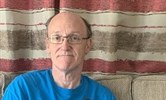 Avatar of Mark Bailey, an HCA with Parkinson's. He is sitting on a sofa looking at the camera, wearing a Parkinson's UK t-shirt