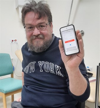 Mark Davies with his controller that is used for the remote reprogramming clinic. Image shows Mark,who is sitting in a wheelchair in a clinic, holding an ipod device