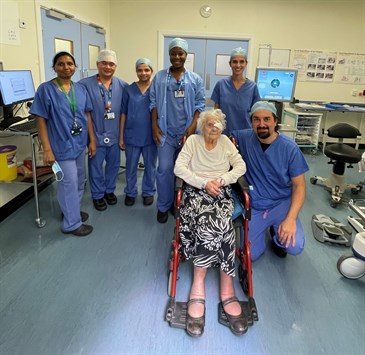 Mary Teal with the surgical team who performed her cataract surgery. Image shows Mary in a wheelchair with a patch over one eye. The surgeon is kneeling next to her and the rest of the team are standing behind her. They are in an operating theatre