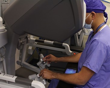 Milan Thomas operating the robot. Image shows Milan in surgical scrubs sitting at the robotic console looking through the viewing device. His hands are on the controls of the robot.