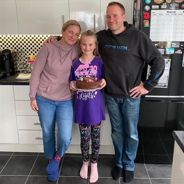 Molly with mum Amy and dad Adam. Image shows them standing in a kitchen, Molly is holding a birthday cake