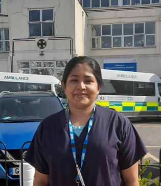 Nav Somasinghe, ST7 trainee. Image shows Nav standing outside the Kent and Canterbury Hospital. There are ambulances and other vehicles in the background. She is wearing scrubs.