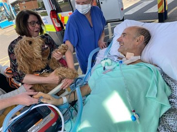Nigel Avery is reunited with Martha the dog. Image shows NIgel in a hospital bed, with ventilator attached, smiling at his dog, who is being held by his wife.  A nurse is looking on. They are pictured outside a hospital.