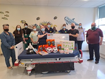 Padua Ward donations. Photo shows people standing behind a hospital bed which is piled high with computer games and devices. Two of the people are holding a giant cheque. They are pictured in a hospital ward.