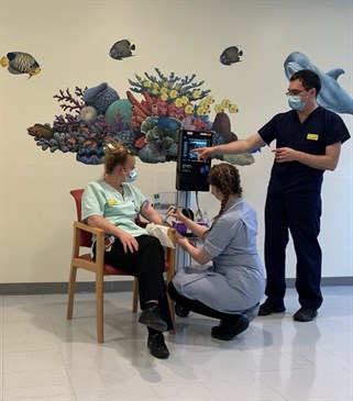 Avatar of Padua ward staff using the vein-finding ultrasound. Photo shows a nurse sitting on a chair while another uses the machine on her arm. A third is standing by the machine.