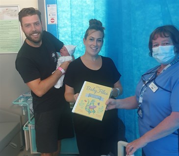 Phil Parrett with Laura and James Willard and baby George. Phil is in uniform and a mask, and Laura is holding one of the books. James is holding the baby and they are pictured in a ward cubicle with blue curtain behind.