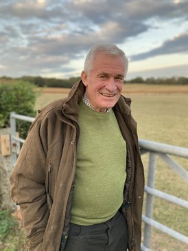 Pip Collick, wearing a green jumper and brown jacket, on his farm. He is looking at the camera with fields behind him.