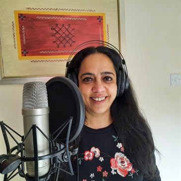 Rema Iyer, a consultant at the QEQM who has recorded a song about a humble rice dish. She is pictured by a professional-looking microphone and is wearing headphones.