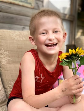 Rosa-Mae Linch, who is receiving treatment for brain tumours on Padua Ward. She is pictured smiling at the camera, holding a sunflower. She is sitting outside wearing a red top and has very little hair.