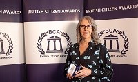 Avatar of Sandy Davies, who was presented with The British Citizen Award. Image shows her holding the award in front of a BCA backdrop