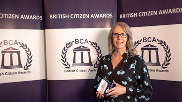Sandy Davies, who was presented with The British Citizen Award. Image shows her holding the award in front of a BCA backdrop