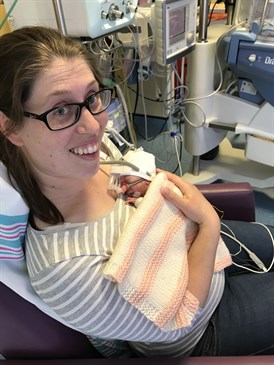 Anna Slark and daughter Amber when she was born. Anna is sitting in a chair, looking up at the camera, holding Amber on her chest. Amber is covered by a blanket, wearing a hat and has a mask over her face.