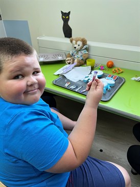 Finley with Rabbit Ray. Image shows him sitting at a table. The toy is on a tray on the table with some tubes of 'blood'. He is holding up one of the tubes and looking back at the camera.