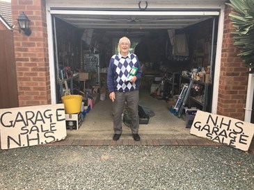 Gerry Collis standing in his garage with items he sells to raise money for East Kent Hospitals Charity. There are two signs saying NHS garage sale