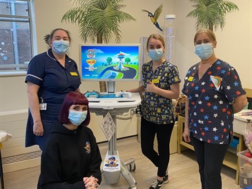 Staff from Padua ward with Jessica Miree from TheRockinR with the games console. The console is on a movable trolley and the staff are standing around it, with Jessica kneeling in front - she is wearing a t-shirt with the charity's logo on it