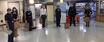 Staff standing in the reception area of the William Harvey Hospital, near the Friends shop. They are standing two metres apart, wearing masks, and holding paper bags containing the food donations