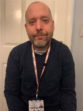 Steve Brown, chair of the disabled staff council. He is sitting in a room in front of a white door with a rainbow lanyard round his neck.